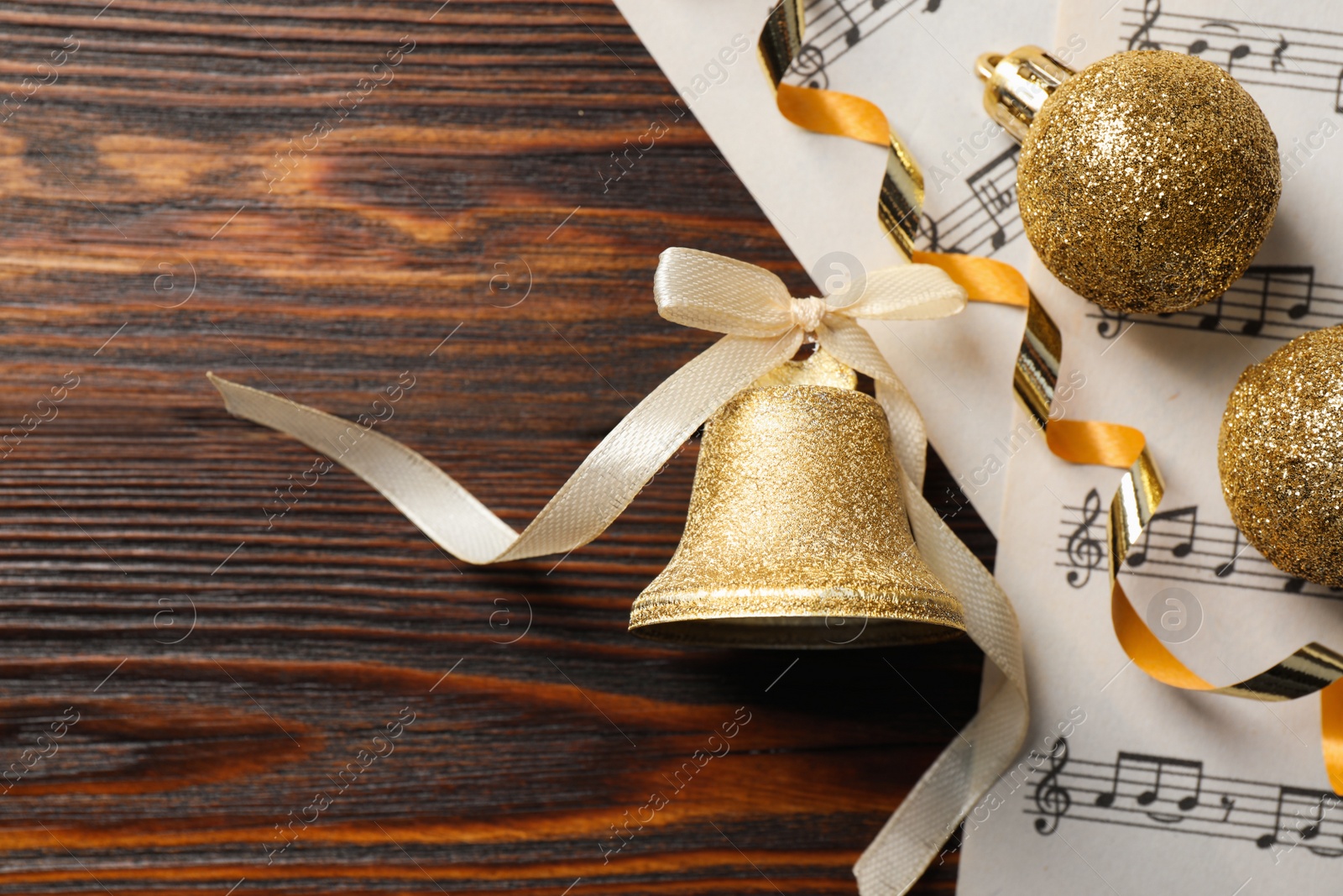 Photo of Golden shiny bell with bow, music sheets and decorative balls on wooden table, flat lay. Christmas decoration