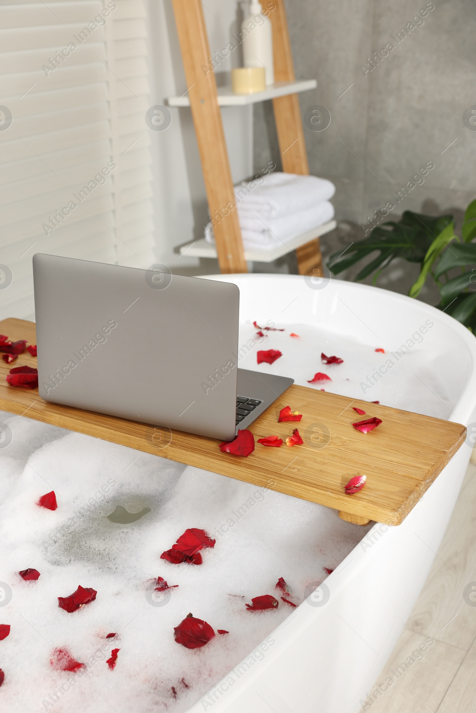 Photo of Wooden board with laptop and rose petals on bath tub in bathroom