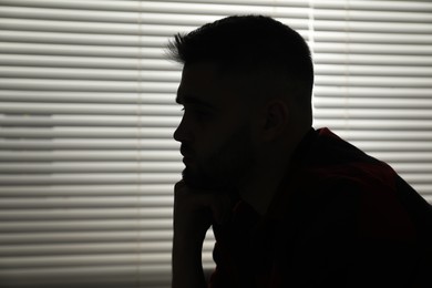 Photo of Silhouette of sad man near closed blinds indoors. Space for text