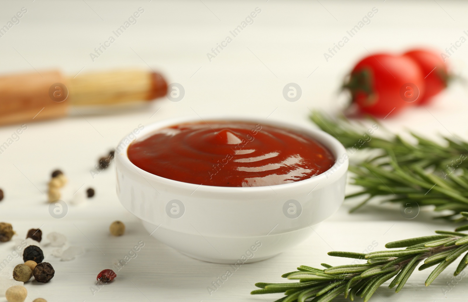 Photo of Tasty barbeque sauce in bowl, rosemary and spices on white wooden table, closeup