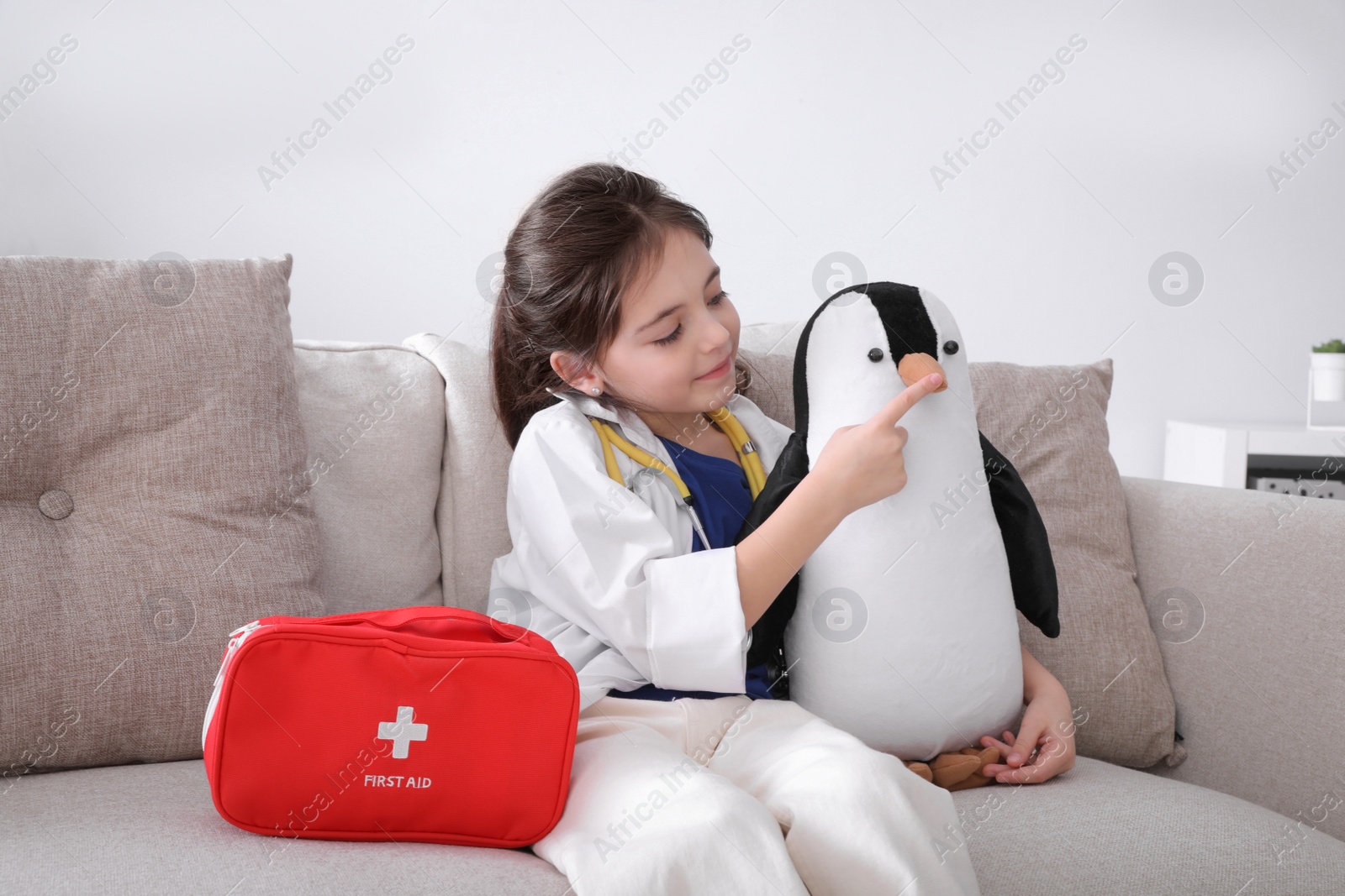 Photo of Little girl playing doctor with toy penguin on sofa indoors. Pediatrician practice