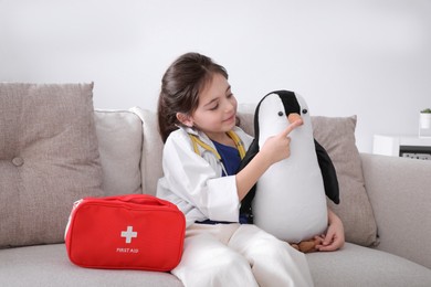 Little girl playing doctor with toy penguin on sofa indoors. Pediatrician practice