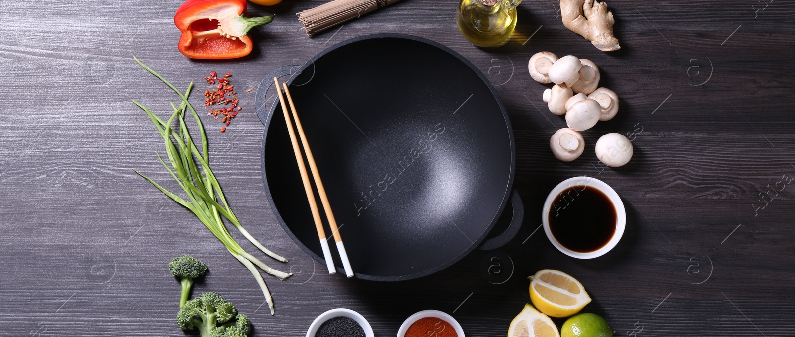 Photo of Empty iron wok and chopsticks surrounded by ingredients on dark grey wooden table, flat lay