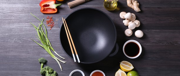 Photo of Empty iron wok and chopsticks surrounded by ingredients on dark grey wooden table, flat lay