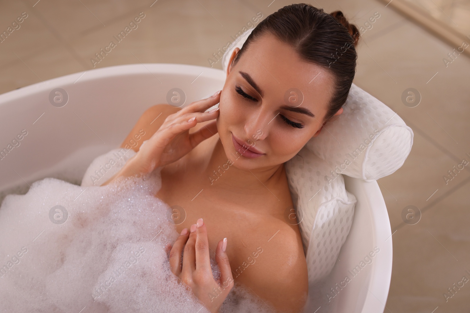 Photo of Young woman using pillow while enjoying bubble bath indoors