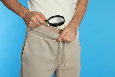 Photo of Man examining intimate area with magnifying glass on light blue background, closeup. Genital herpes