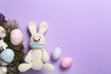 Photo of Toy bunny with protective mask, painted eggs and space for text on lilac background, flat lay. Easter holiday during COVID-19 quarantine