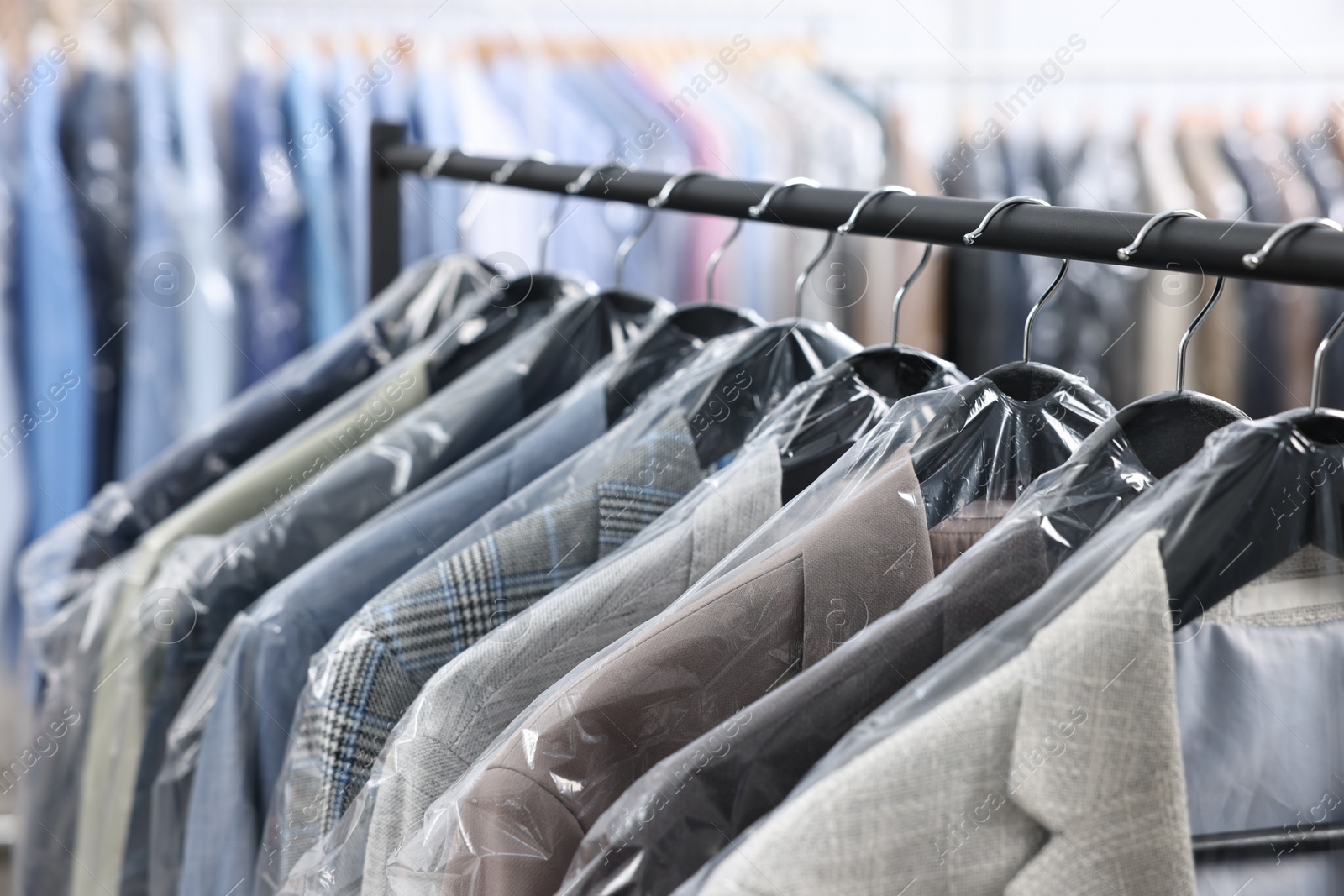 Photo of Dry-cleaning service. Many different clothes in plastic bags hanging on rack indoors, closeup