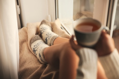 Photo of Woman in knitted socks holding cup of tea on plaid near window at home, closeup