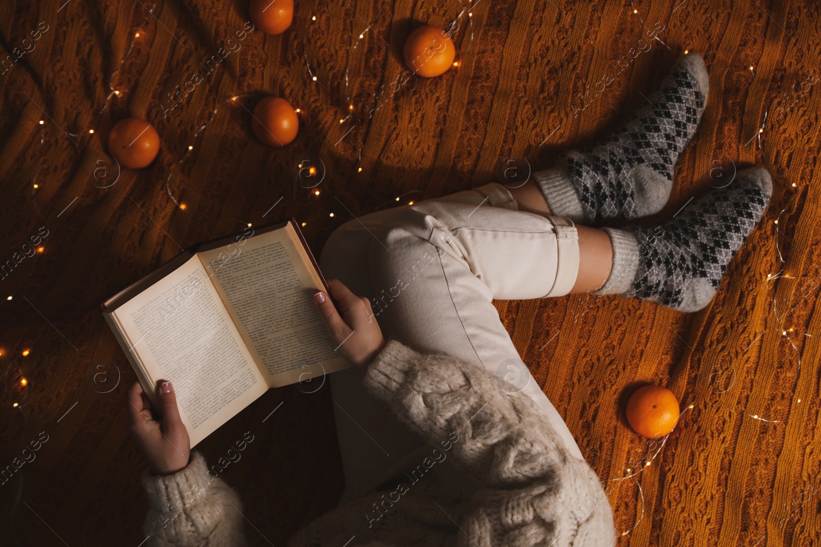 Photo of Woman in warm socks with book relaxing on knitted blanket, top view