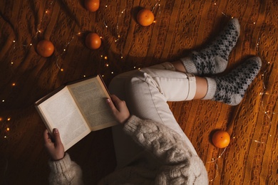 Woman in warm socks with book relaxing on knitted blanket, top view