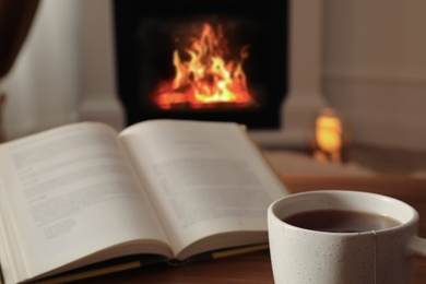 Photo of Cup of hot tea and book on wooden table near fireplace at home. Cozy atmosphere