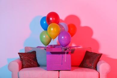 Photo of Gift box with bright air balloons on sofa against color background