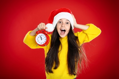 Girl in Santa hat with alarm clock on red background. New Year countdown