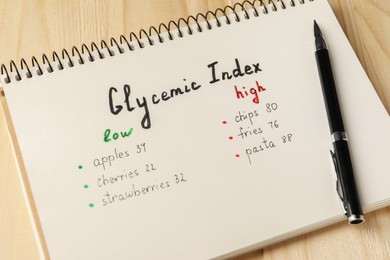 List with products of low and high glycemic index in notebook and pen on wooden table, closeup