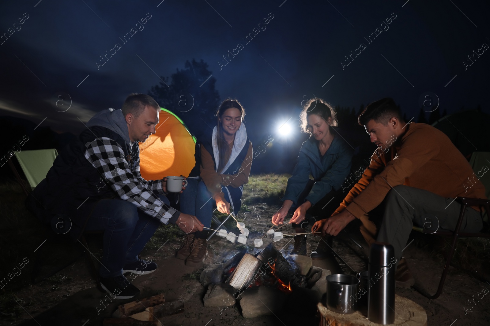 Photo of Group of friends roasting marshmallows on bonfire at camping site in evening