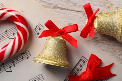 Photo of Golden shiny bells with red bows, music sheets and candy cane on wooden table, flat lay. Christmas decoration