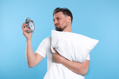 Photo of Unhappy man with pillow and alarm clock on light blue background. Insomnia problem