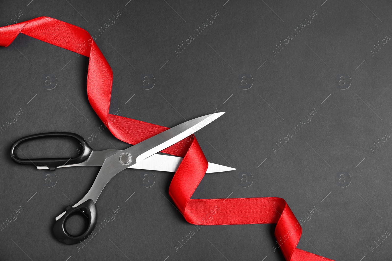 Photo of Stylish scissors and red ribbon on black background, flat lay with space for text. Ceremonial tape cutting