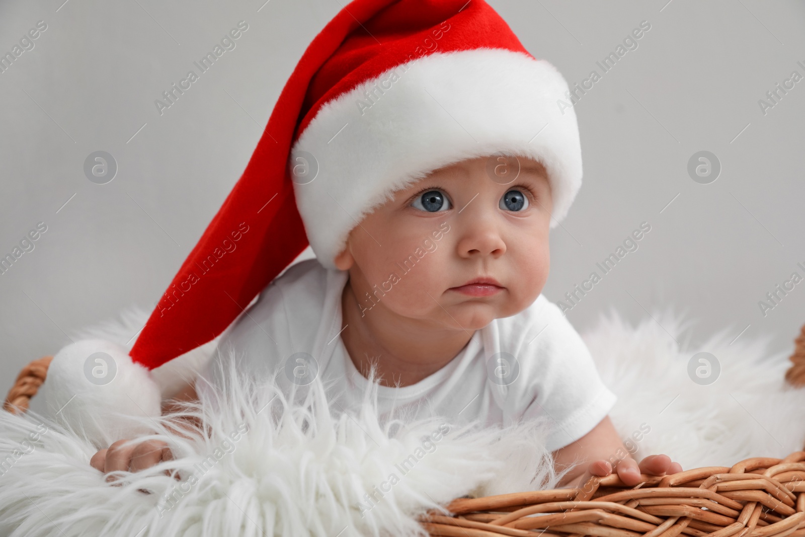 Photo of Cute baby wearing Santa hat in wicker basket on light grey background. Christmas celebration