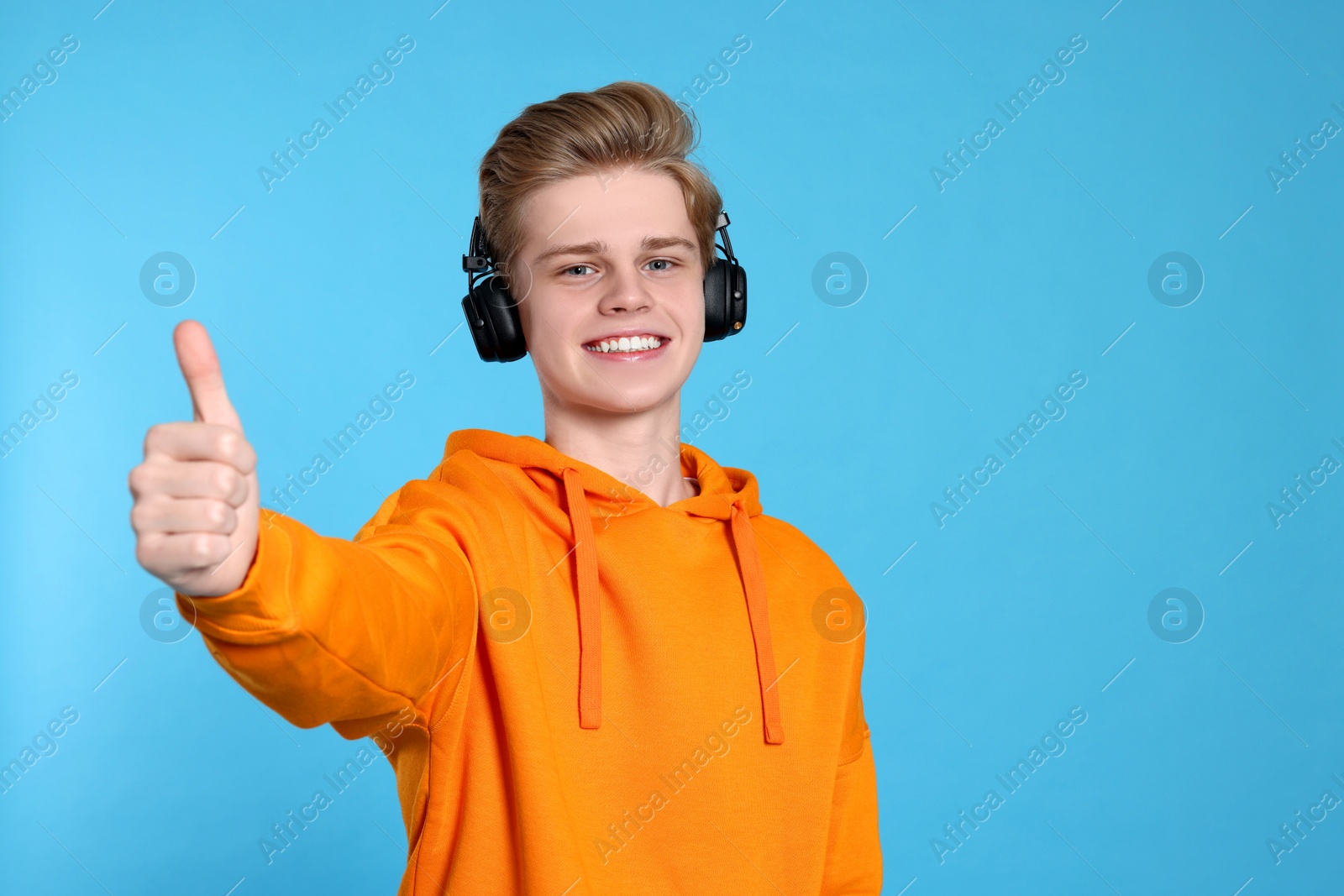 Photo of Teenage boy with headphones showing thumb up on light blue background. Space for text