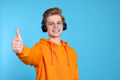 Teenage boy with headphones showing thumb up on light blue background. Space for text