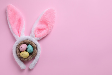 Headband with Easter bunny ears and dyed eggs in nest on pink background, flat lay. Space for text