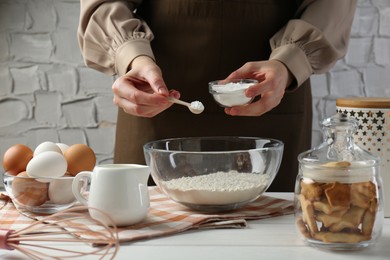 Photo of Woman with spoon and bowl of baking powder at white wooden table, closeup