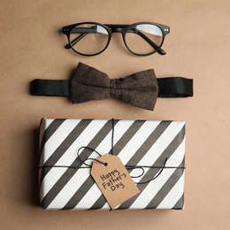 Photo of Eyeglasses, bow tie and gift with tag HAPPY FATHER'S DAY on brown background, flat lay