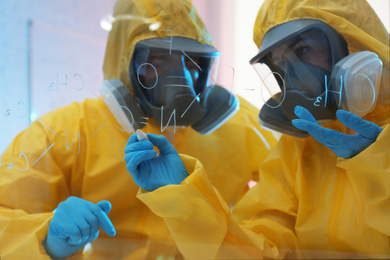 Scientists in chemical protective suits writing formula on glass board at laboratory. Virus research
