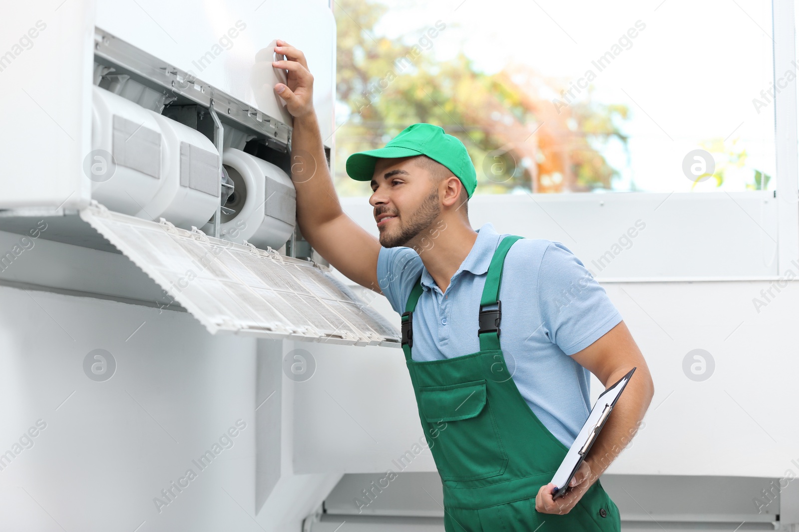 Photo of Professional technician with clipboard maintaining modern air conditioner indoors