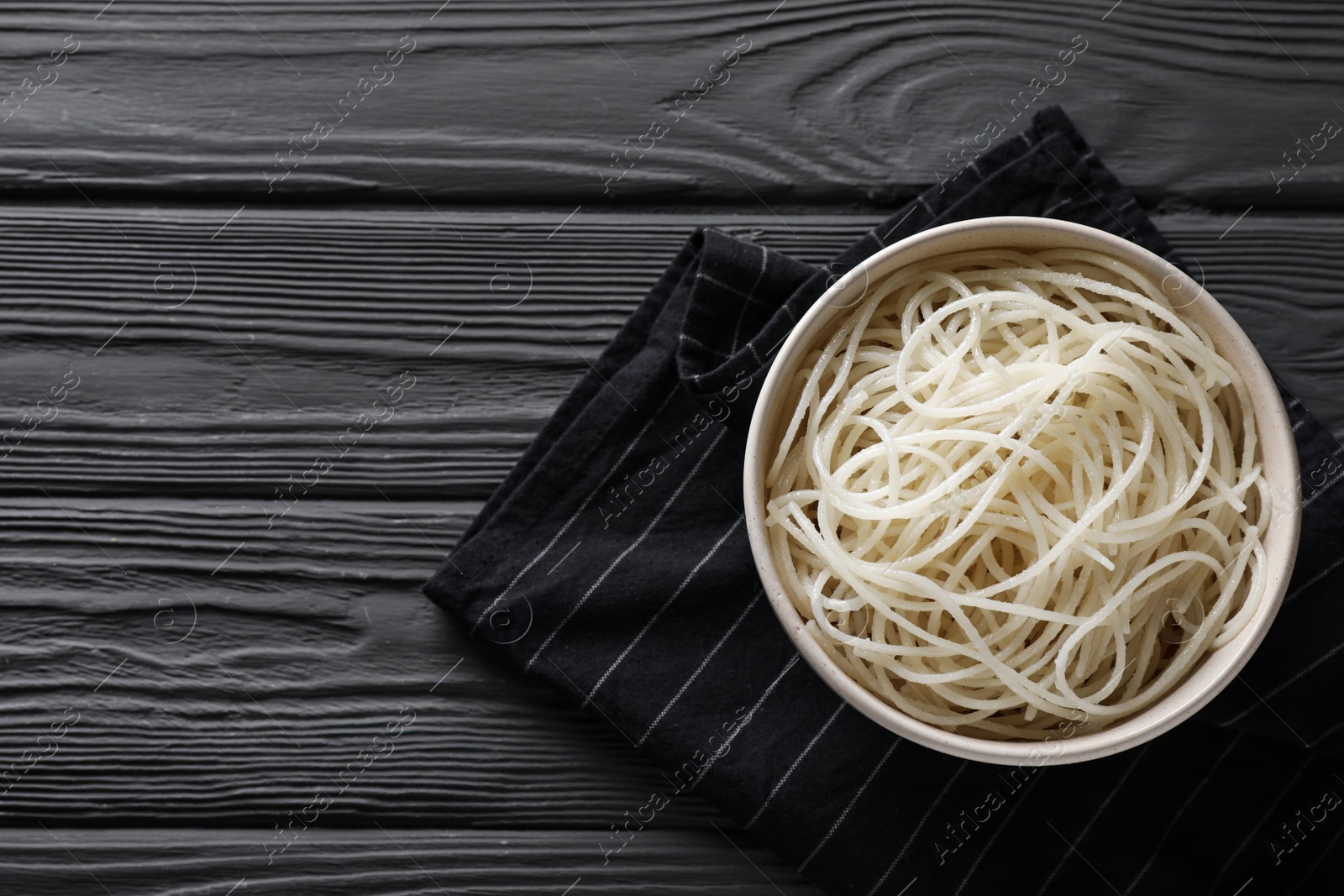 Photo of Bowl of tasty cooked rice noodles and napkin on black wooden table, top view. Space for text