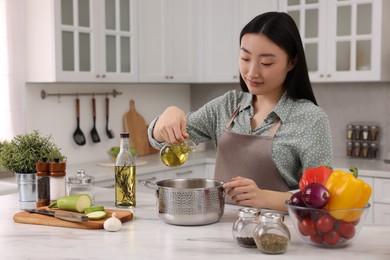 Cooking process. Beautiful woman pouring oil from bottle into pot at white countertop in kitchen