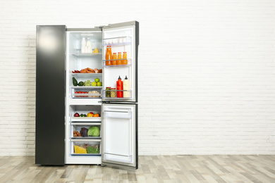 Photo of Open refrigerator filled with food near white brick wall, space for text