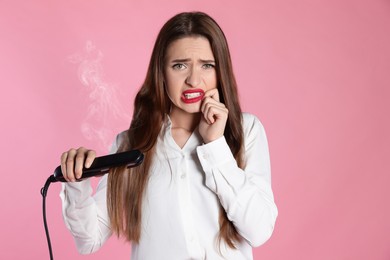 Photo of Upset young woman with flattening iron on light pink background. Hair damage