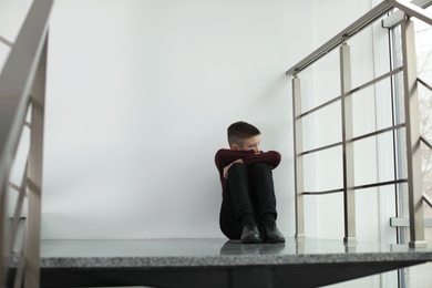 Photo of Upset boy sitting on staircase indoors. Space for text