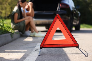 Photo of Couple sitting near broken car on roadside outdoors, focus on warning triangle
