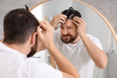Photo of Dandruff problem. Man with comb examining his hair and scalp near mirror indoors