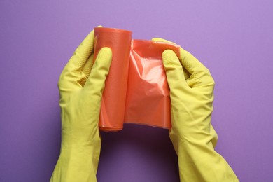 Janitor in rubber gloves holding roll of orange garbage bags over purple background, top view