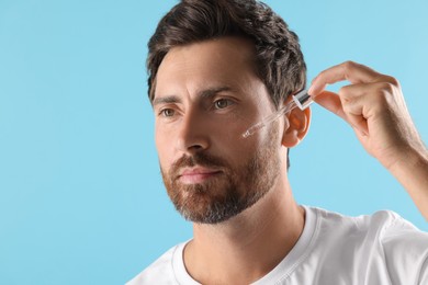 Handsome man applying cosmetic serum onto his face on light blue background. Space for text