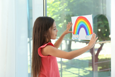 Photo of Little girl with picture of rainbow near window indoors.  Stay at home concept