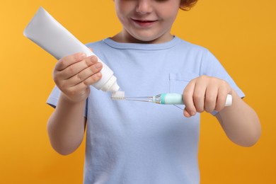 Photo of Cute little boy squeezing toothpaste from tube onto electric toothbrush on yellow background, closeup