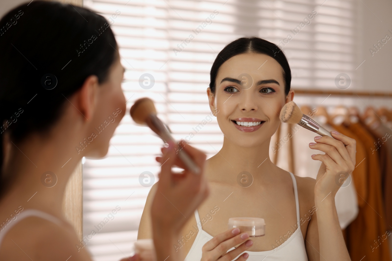 Photo of Beautiful young woman applying face powder with brush in front of mirror at home