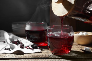Pouring delicious hibiscus tea from glass teapot into cup at wooden table, closeup