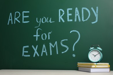 Photo of Books and alarm clock on wooden table near chalkboard with phrase Are You Ready For Exams