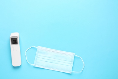 Photo of Infrared thermometer and medical mask on light blue background, flat lay with space for text. Checking temperature during Covid-19 pandemic