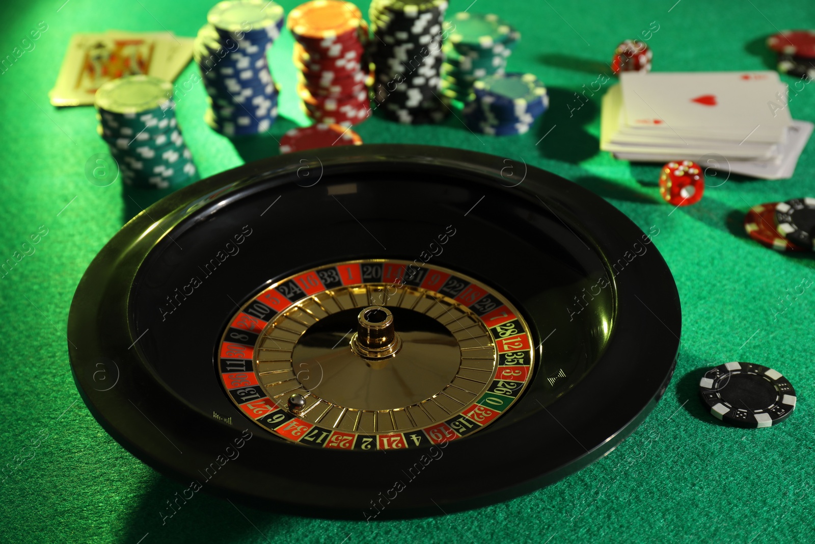 Photo of Roulette wheel with ball, playing cards and chips on green table. Casino game