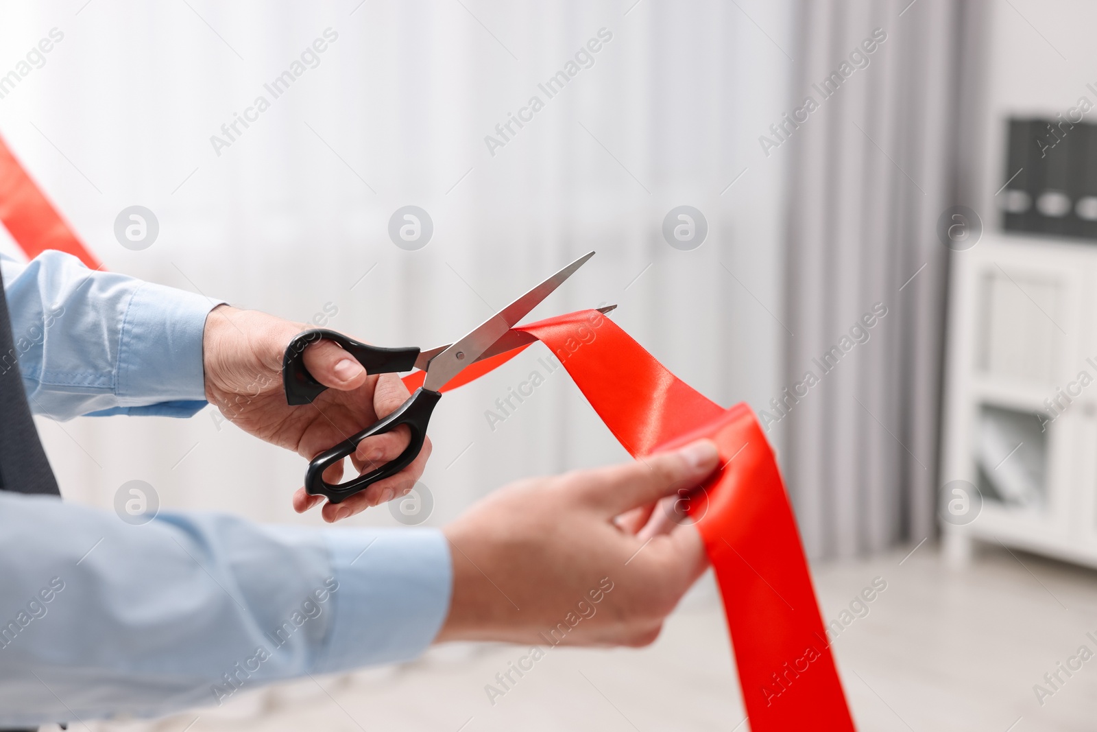 Photo of Man cutting red ribbon with scissors indoors, closeup