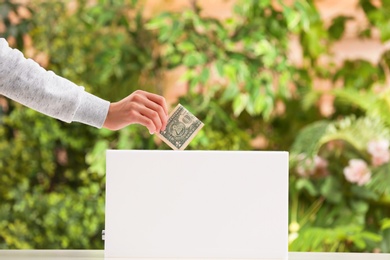 Photo of Woman putting money into donation box on blurred background, closeup. Space for design