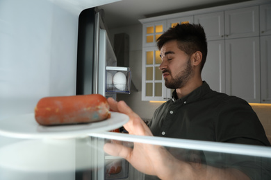 Young man feeling bad smell of spoiled sausage in refrigerator, view from inside